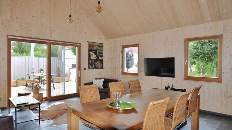 Ein modernes Wohnzimmer mit einem großen Esstisch und hellen Holzdetails. Große Fenster bieten einen Blick auf den Außenbereich.