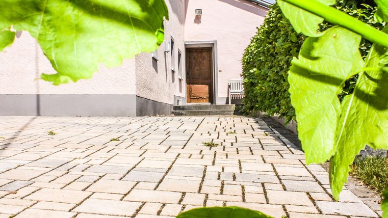 Ein gepflasterter Weg führt zur Haustür eines Hauses. Auf der linken Seite sind grüne Blätter sichtbar.