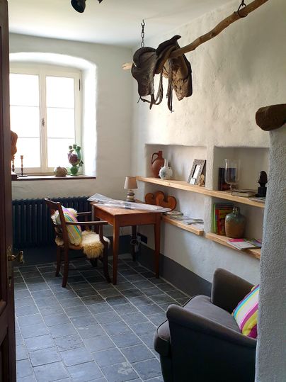 Een gezellige ruimte met een bureau, een comfortabele stoel en planken. Aan de muur hangen enkele hoeden, en een raam zorgt voor daglicht.