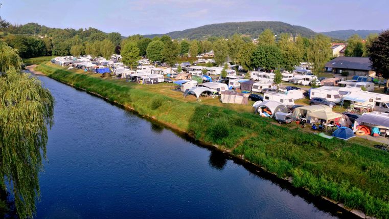 Un camping au bord de la rivière avec de nombreuses caravanes et tentes. Entouré d'arbres et de douces collines, le paysage offre une atmosphère calme.