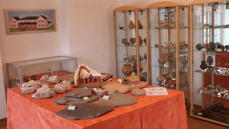 Ein Ausstellungsraum mit einer roten Tischdecke, auf dem verschiedene Gegenstände zu sehen sind. In Vitrinen sind historische Artefakte und Keramiken ausgestellt.