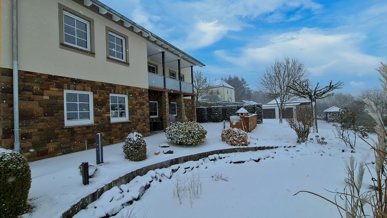 Une maison dans la neige avec un jardin bien entretenu. Le ciel est bleu avec quelques nuages.