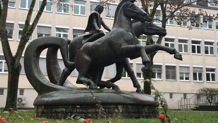 Eine bronzeartige Statue eines Reiters auf einem Pferd steht in einem Grünbereich vor einem Gebäude. Die Statue zeigt den Reiter in ruhiger Haltung mit erhobenem Schwert.
