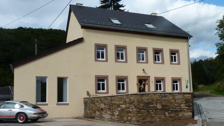 Ein modernes, zweigeschossiges Wohngebäude mit hellgelber Fassade und grauem Dach. Vor dem Haus stehen einige Personen und ein silbernes Auto parkt auf dem Grundstück.