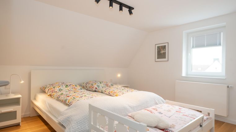 Une chambre claire avec un grand lit et un lit d'enfant supplémentaire.
Les murs sont clairs, et une fenêtre laisse entrer beaucoup de lumière du jour.