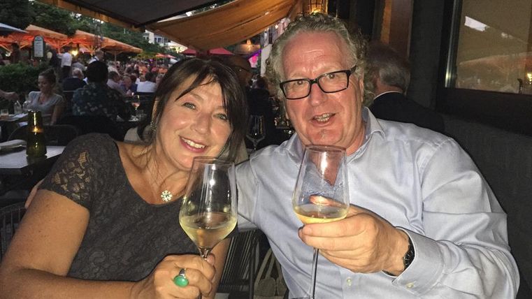 Ein fröhliches Paar sitzt bei Kerzenschein in einem Restaurant und stößt mit Weingläsern an. Im Hintergrund sind weitere Gäste und eine gemütliche Atmosphäre zu sehen.