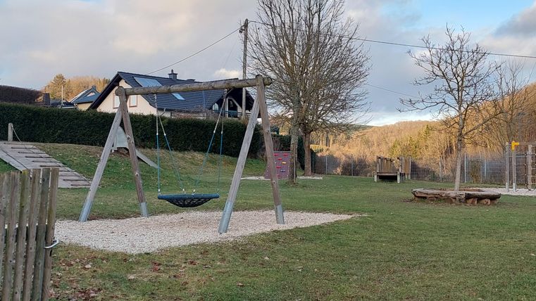 A playground with a swing and a seesaw. In the background, there are trees and a house.