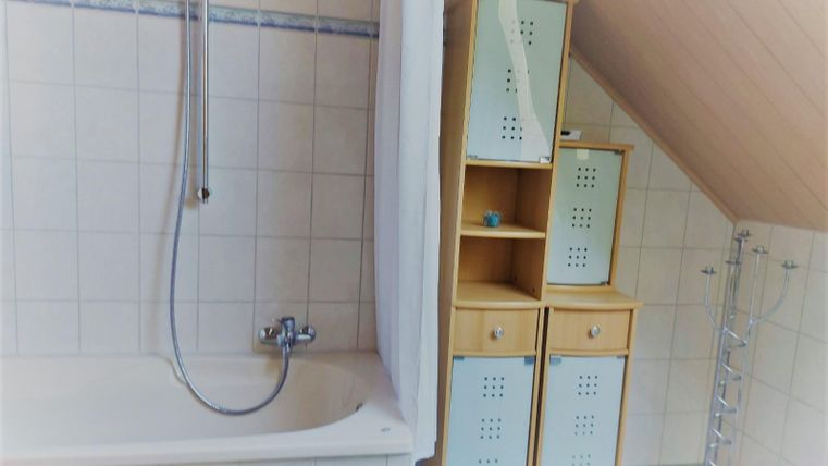 Een badkamer met een bad en een douche-installatie. Naast het bad staat een helder rek met vakken voor opslag.