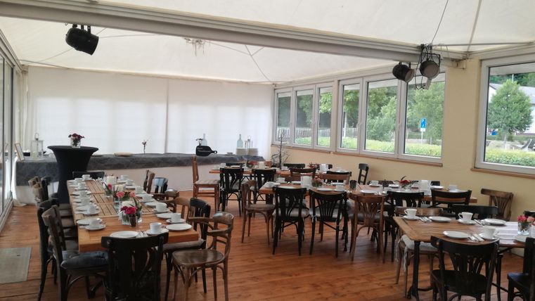 An elegant event space with wooden tables and chairs. Light streams through large windows, and the room is ready for an event.