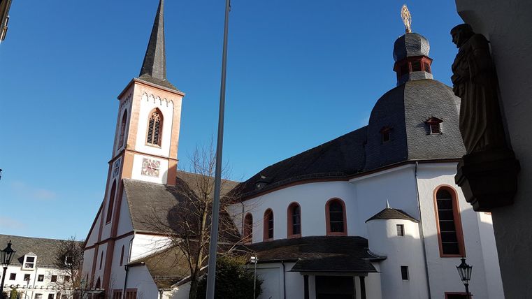 Eine Kirche mit einem hohen Turm und einer markanten Kuppel auf der rechten Seite. Der Himmel ist klar und blau, umgeben von einer ruhigen Stadtansicht.