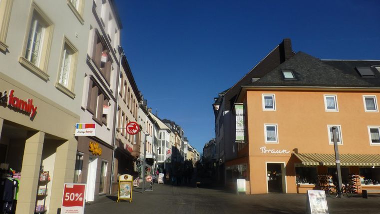 Eine ruhige Einkaufsstraße mit bunten Gebäuden und blauem Himmel. Auf der linken Seite sind Geschäfte und Werbeplakate sichtbar.