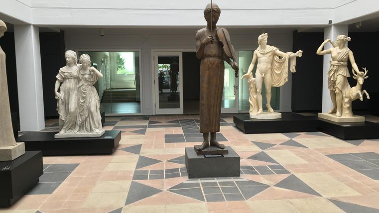 An interior view with several sculptures on pedestals. The room has a modern floor and large windows that let in light.