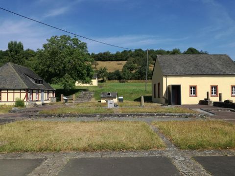 Eine ruhige Landschaft mit traditionellen Gebäuden und einer großen, grünen Wiese. Im Hintergrund sind sanfte Hügel und ein klarer Himmel zu sehen.