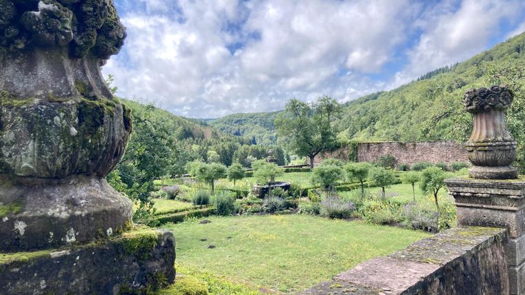 Eine wunderschöne Gartenlandschaft mit grünen Wiesen und Bäumen. Im Hintergrund sind sanfte Hügel und ein klarer Himmel zu sehen.
