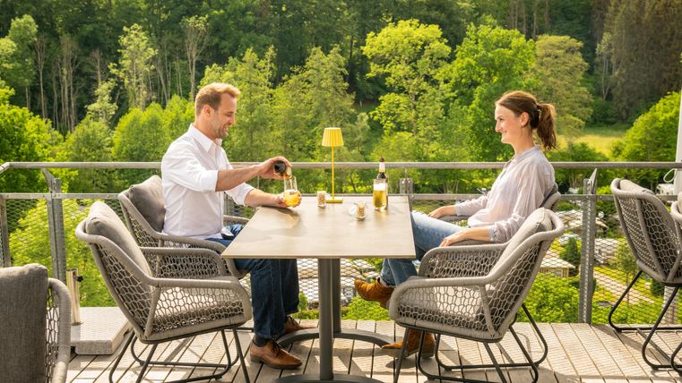 Un couple est assis à une table sur une terrasse et déguste des boissons. En arrière-plan, on peut voir des arbres verts et un beau paysage.