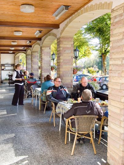 Une terrasse confortable avec plusieurs clients qui profitent de leurs repas. Une serveuse sert des plats sous des arbres ombragés.