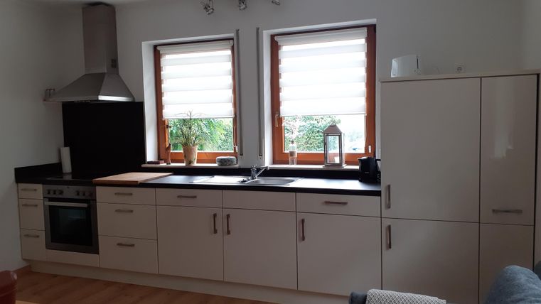 Une cuisine moderne avec des meubles clairs et un plan de travail en bois. Deux fenêtres laissent entrer beaucoup de lumière du jour et une plante sert de décoration.