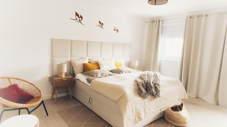Ein helles Schlafzimmer mit einem großen Bett und dekorativen Kissen. Es gibt einen gemütlichen Stuhl und viel Tageslicht durch das Fenster.