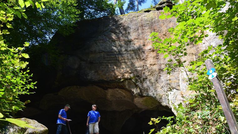 Een natuurlijke rotsformatie in het bos met twee personen ervoor. Omringd door weelderig groen en bomen.