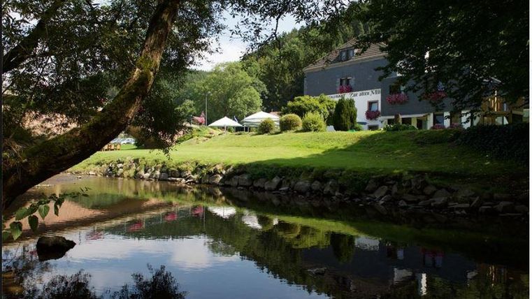 Un paysage tranquille avec une rivière claire, entourée d'arbres. En arrière-plan, on voit un bâtiment et des prairies verdoyantes.