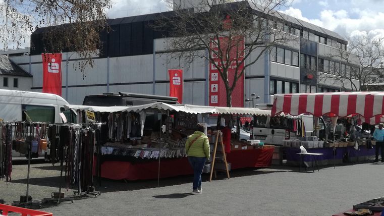 Ein Markt mit verschiedenen Ständen und Verkaufsflächen. Im Hintergrund sind Gebäude und Bäume zu sehen.