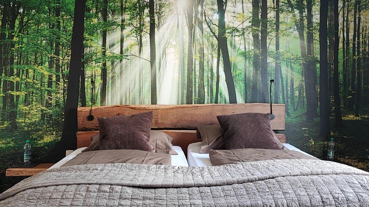 Ein gemütliches Schlafzimmer mit einem Holzbett und weichen Kissen. Die Wand zeigt einen schönen Wald mit Sonnenlicht, das durch die Bäume scheint.