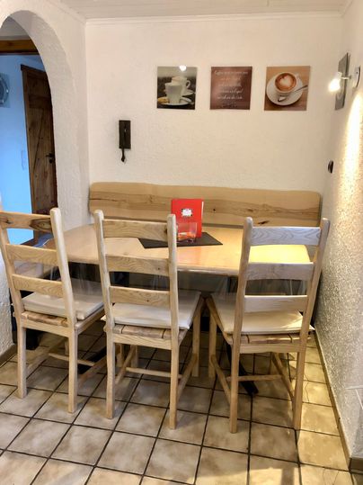 A cozy dining area with a wooden table, three chairs, and a corner bench. On the wall hang some pictures, and there is some decor on the table.