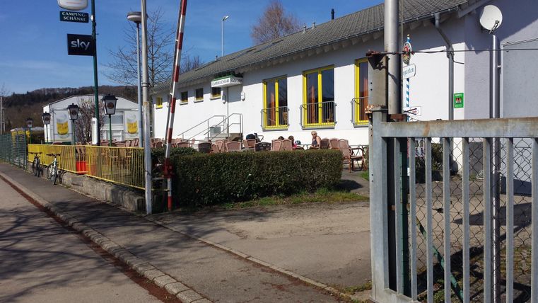 Ein Gebäude mit weißen Wänden und großen Fenstern, umgeben von einem gepflegten Garten. Vor dem Gebäude befinden sich Sitzgelegenheiten im Freien.