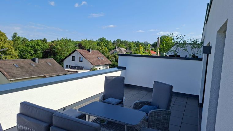 A modern rooftop terrace with seating and a view of green trees and houses. The sky is clear and sunny.