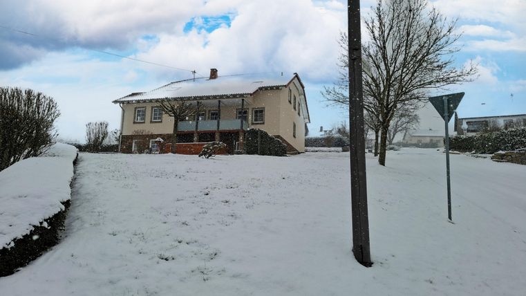 Une maison dans la neige avec un paysage enneigé. Le ciel est nuageux et il y a quelques arbres dans les environs.