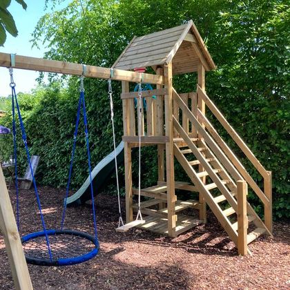 Un terrain de jeux avec un toboggan en bois, une excavation de sable et des balançoires. L'environnement est entouré de plantes vertes.