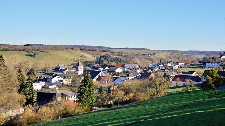 Blick auf ein Dorf in der Südeifel mit grünen Wiesen und blauen Himmel.