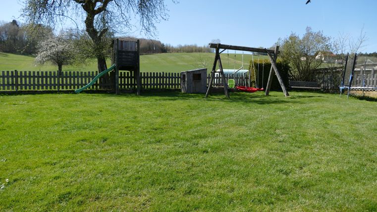 A green garden with a swing and a playhouse. In the background, gentle hills and a blue sky can be seen.
