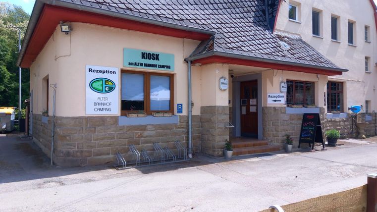 Een ontvangstgebouw met een kiosk op een camping. De gevel is eenvoudig en uitnodigend, omgeven door een parkeerplaats.