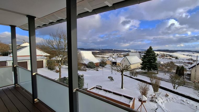 Ein Blick von einem Balkon auf eine verschneite Landschaft. Im Hintergrund sind Häuser und Bäume sichtbar, und der Himmel ist teilweise bewölkt.
