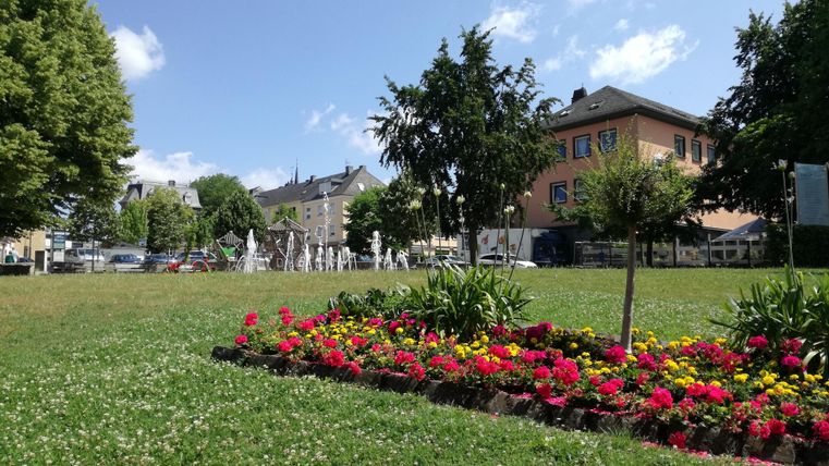 Ein schöner Park mit bunten Blumenbeeten und grüner Wiese. Im Hintergrund sind Bäume und ein Wasserbrunnen zu sehen.