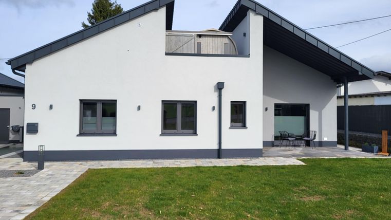 Une maison moderne de plain-pied avec un toit gris et de grandes fenêtres. Le jardin est vert et bien entretenu, avec une zone pavée devant la maison.