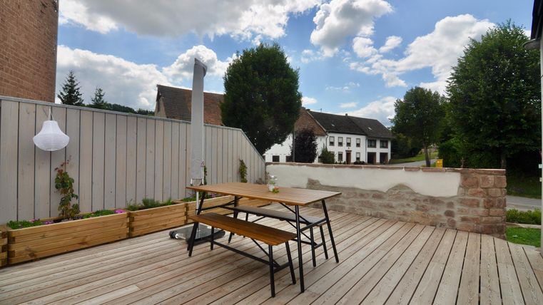 Eine gemütliche Terrasse mit Holzfußboden und einem Tisch. Im Hintergrund sind Bäume und ein Gebäude sichtbar.