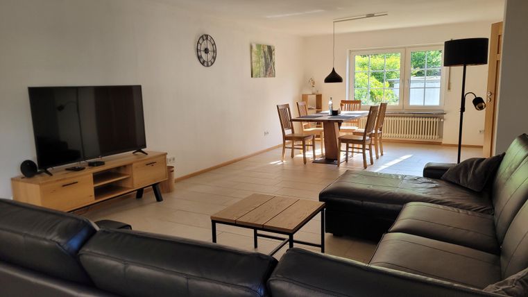 Ein modernes Wohnzimmer mit einem großen Fernseher, einer komfortablen Couch und einem Essbereich. Es hat viele Fenster, die natürliches Licht hereinlassen.