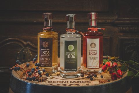 Drei Flaschen Spirituosen stehen auf einem Holzfass. Jede Flasche hat ein anderes Etikett und ist umgeben von dekorativen Naturmaterialien.