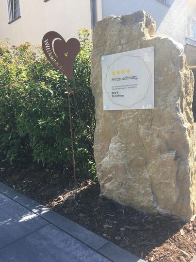 Une grande pierre avec un panneau de bienvenue et un symbole de cœur se trouve dans un paysage de jardin. L'environnement est vert et amical.