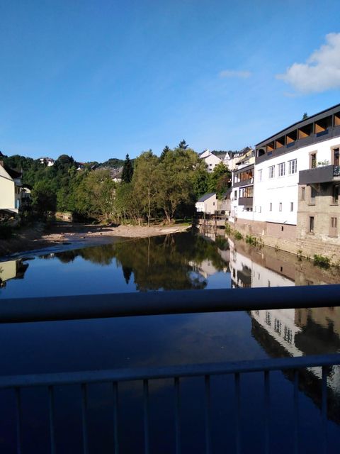 Une rivière calme avec des reflets des bâtiments et des arbres sur la rive. Le ciel est clair et bleu, ce qui crée une atmosphère paisible.
