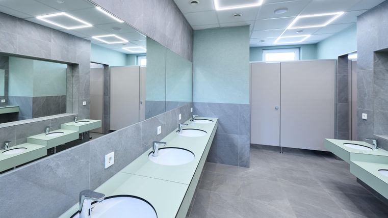 Ein modernes Badezimmer mit mehreren Waschbecken und großen Spiegeln. Die Wände sind in neutralen Farbtönen gehalten, und es gibt geschlossene Kabinen für die Privatsphäre.