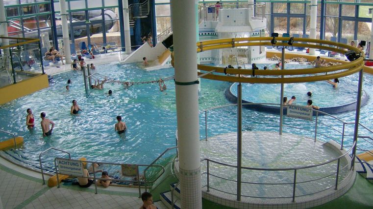 Ein großes Hallenbad mit verschiedenen Wasserbereichen und einer Rutsche. Viele Menschen genießen das Schwimmen und Spielen im Wasser.