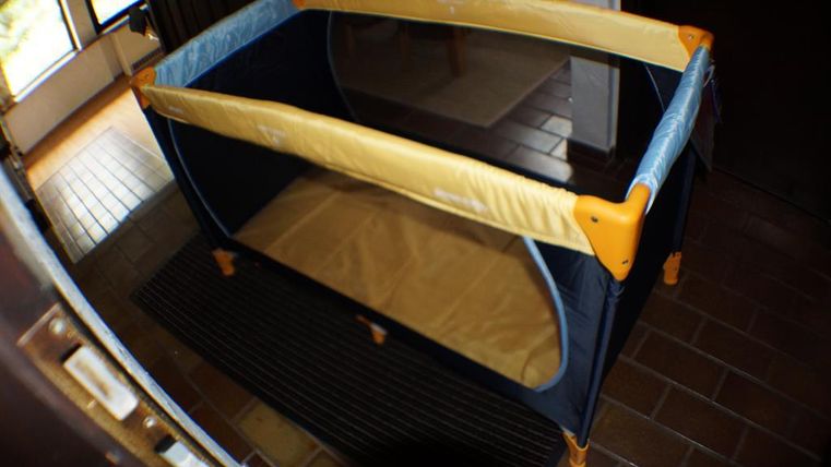 Un lit de voyage dans une pièce, avec des éléments bleus et jaunes. Le sol est en carreaux de bois et il y a de la lumière naturelle provenant d'une fenêtre.