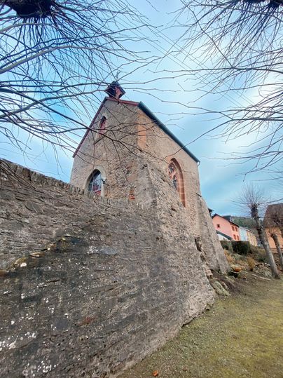 Eine historische Kirche steht in der Nähe einer Steinmauer. Der Himmel ist bewölkt und die Umgebung wirkt ruhig.