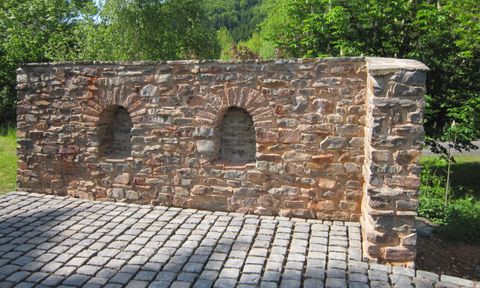 Eine alte Steinmauer mit zwei Bogenfenstern. Die Mauer ist von grünen Bäumen umgeben und steht auf einem gepflasterten Boden.