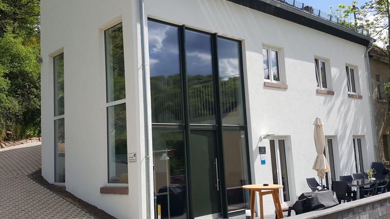 Ein modernes, zweistöckiges Haus mit großen Fenstern und einem klaren Design. Der Außenbereich ist mit Sitzgelegenheiten ausgestattet und von einer grünen Umgebung umgeben.