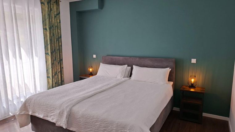 A modern bedroom with a double bed and a green wall. Large windows provide plenty of daylight and a cozy atmosphere.