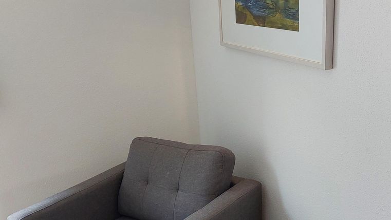 Een gezellige fauteuil staat in de hoek van een kamer. Naast de fauteuil staat een kleine houten tafel en aan de muur hangt een afbeelding.